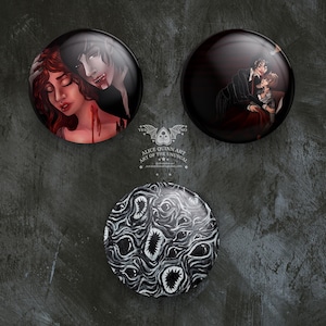 May include: Three round, glossy buttons with dark, gothic-themed illustrations. One depicts a vampire and a woman, another a couple embracing, and the third a black and white pattern of eyes and mouths. The buttons are arranged on a textured, dark gray surface.