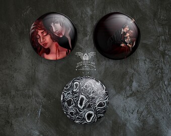 May include: Three round, glossy buttons with dark, gothic-themed illustrations. One depicts a vampire and a woman, another a couple embracing, and the third a black and white pattern of eyes and mouths. The buttons are arranged on a textured, dark gray surface.