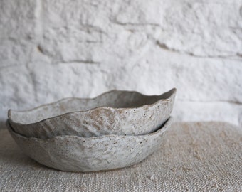 May include: Two white ceramic bowls with a textured, uneven surface. The bowls are stacked on top of each other and are sitting on a light brown fabric surface.