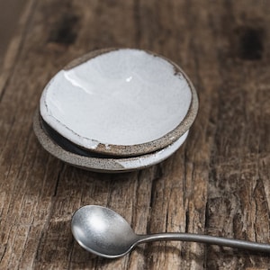 Op de afbeelding: Een witte keramische kom met een bruine rand staat op een houten oppervlak. Een zilveren lepel ligt naast de kom.