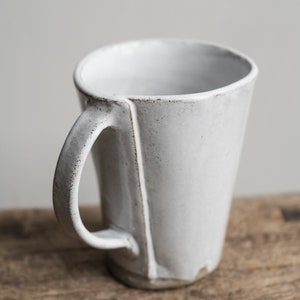 May include: A white ceramic mug with a unique handle and a speckled glaze. The mug is sitting on a rustic wooden surface.