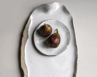 Puede incluir: Una fuente de cerámica blanca con borde irregular, que sostiene un plato más pequeño con dos higos maduros. La fuente tiene una superficie texturizada blanquecina y un borde natural sin esmaltar. Los higos son de color púrpura oscuro.