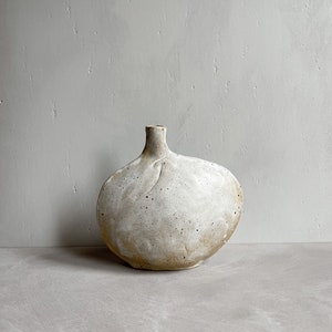 Könnte beinhalten: Eine weiße Keramikvase mit unregelmäßiger, runder Form. Die Vase hat eine strukturierte Oberfläche und eine leicht gebrochenweiße Farbe.