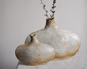 May include: Two ceramic vases in a speckled white and brown glaze. The larger vase is taller and wider than the smaller vase. Both vases have a rounded, organic shape. A dried sprig of flowers is in the larger vase.