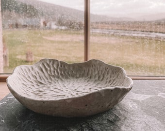 May include: A white ceramic bowl with a textured surface sits on a gray countertop. The bowl has a rounded, organic shape and is slightly chipped on the edge.