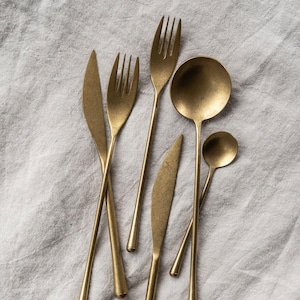 May include: A selection of gold-toned cutlery, comprising forks, knives, and spoons, displayed on a light grey linen cloth. The utensils feature a matte finish and a minimalist design, ideal for a sophisticated dining experience.