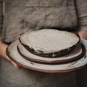 Pu&ograve; includere: Un set di tre piatti in ceramica bianca con accenti marroni. I piatti sono impilati uno sopra l'altro e hanno un aspetto rustico e fatto a mano.