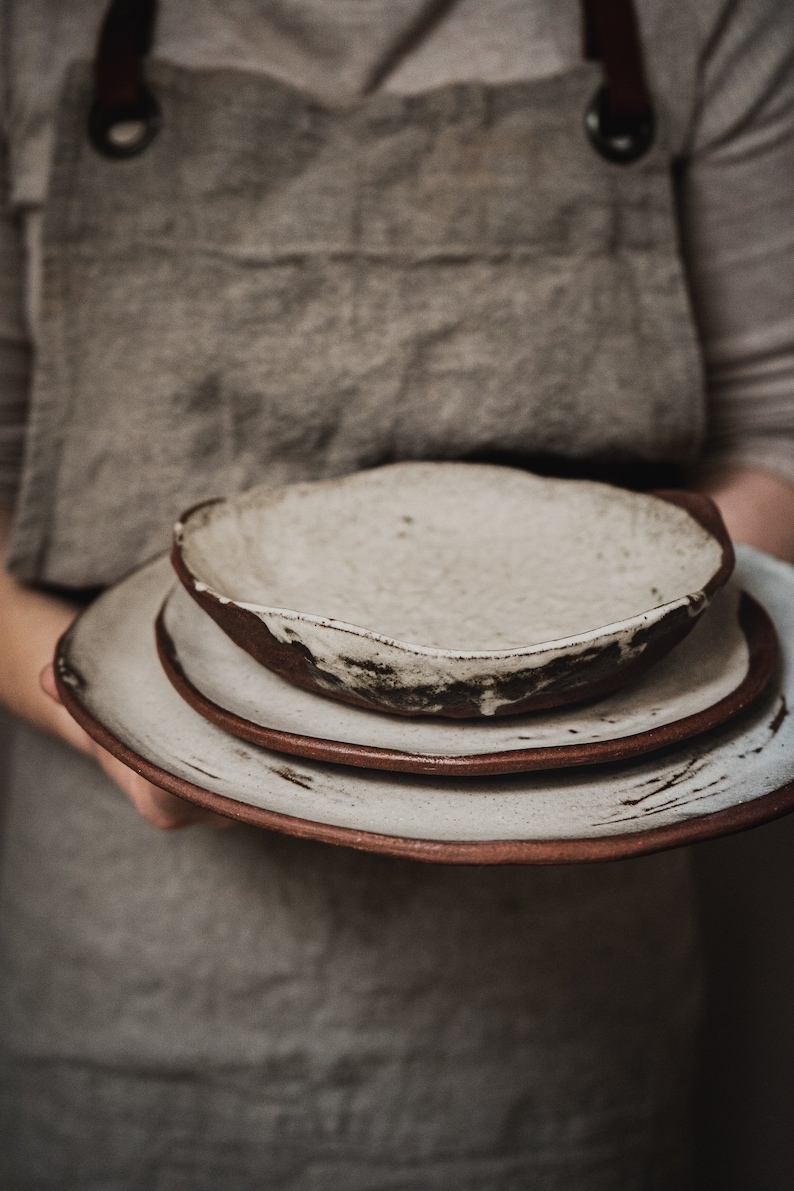 Pu&ograve; includere: Un set di tre piatti in ceramica bianca con accenti marroni. I piatti sono impilati uno sopra l'altro e hanno un aspetto rustico e fatto a mano.
