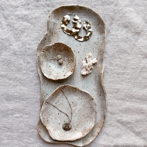 May include: A set of three white ceramic dishes with a speckled glaze. The dishes are arranged in a stack, with the largest dish at the bottom and the smallest dish at the top. The dishes are decorated with gold earrings, a necklace with a sun pendant, and a small white coral piece.