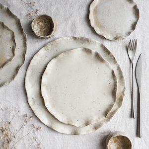 Op de afbeelding: Een verzameling handgemaakt keramisch servies, waaronder borden en kleine kommen, met een gespikkelde, gebroken witte glazuur en een getextureerde, golvende rand. De set bevat een vork en een mes. De rustieke, neutrale tinten cre&euml;ren een natuurlijke, organische esthetiek.