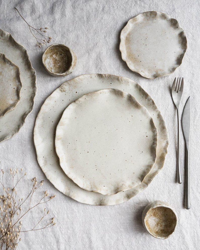 Op de afbeelding: Een verzameling handgemaakt keramisch servies, waaronder borden en kleine kommen, met een gespikkelde, gebroken witte glazuur en een getextureerde, golvende rand. De set bevat een vork en een mes. De rustieke, neutrale tinten cre&euml;ren een natuurlijke, organische esthetiek.