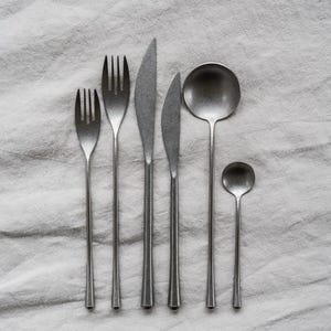 Op de afbeelding: Een set roestvrijstalen bestek, waaronder vorken, messen, een soeplepel en een lepel, gerangschikt op een wit linnen oppervlak. De gebruiksvoorwerpen hebben lange, slanke handvatten en een matte afwerking, wat een minimalistische uitstraling cre&euml;ert.