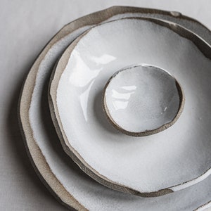 Pu&ograve; includere: Tre piatti in ceramica bianca con un bordo marrone, impilati uno sopra l'altro. I piatti sono di dimensioni diverse, con il piatto pi&ugrave; piccolo al centro.