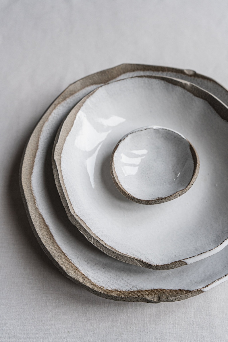 Pu&ograve; includere: Tre piatti in ceramica bianca con un bordo marrone, impilati uno sopra l'altro. I piatti sono di dimensioni diverse, con il piatto pi&ugrave; piccolo al centro.
