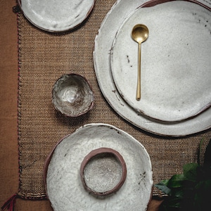 Pu&ograve; includere: Un set di piatti e ciotole in ceramica bianca dall'aspetto rustico e fatto a mano. I piatti sono impilati uno sopra l'altro e le ciotole sono posizionate accanto. Un cucchiaio dorato &egrave; posto sopra i piatti. I piatti e le ciotole sono su una superficie di juta marrone.