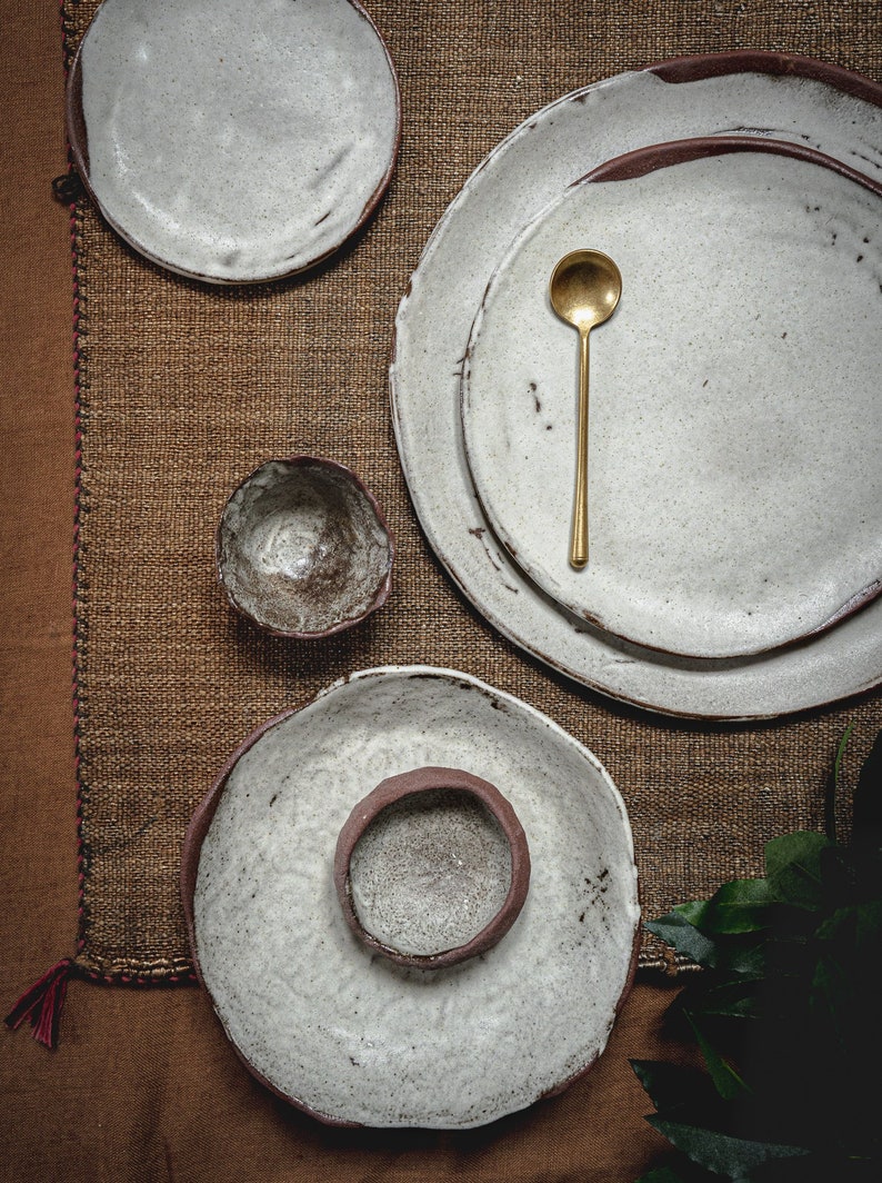 Pu&ograve; includere: Un set di piatti e ciotole in ceramica bianca dall'aspetto rustico e fatto a mano. I piatti sono impilati uno sopra l'altro e le ciotole sono posizionate accanto. Un cucchiaio dorato &egrave; posto sopra i piatti. I piatti e le ciotole sono su una superficie di juta marrone.