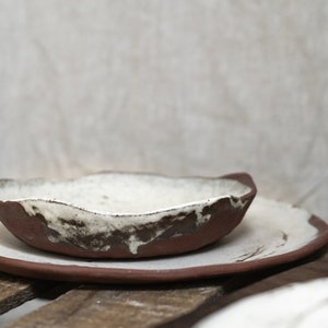 Pu&ograve; includere: Un set di due piatti in ceramica, uno pi&ugrave; piccolo dell'altro, con una glassa bianca e accenti marroni. I piatti sono impilati l'uno sull'altro e sono appoggiati su una superficie di legno.