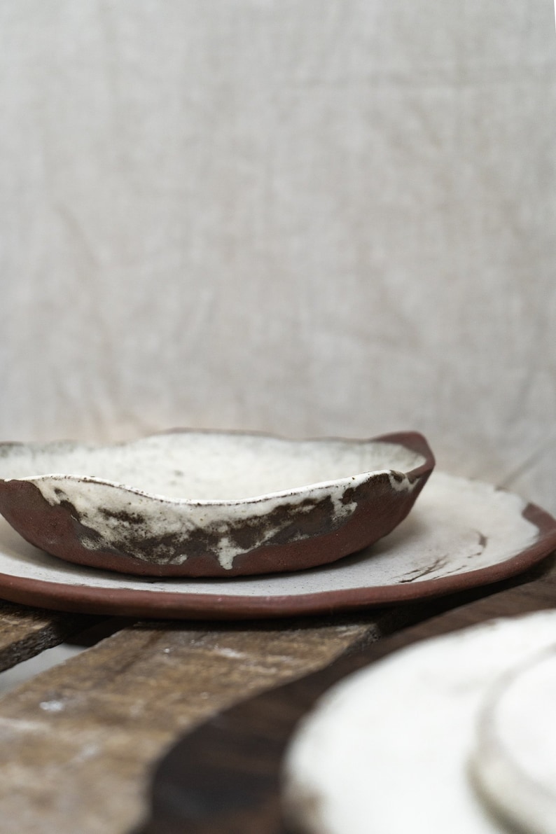 Pu&ograve; includere: Un set di due piatti in ceramica, uno pi&ugrave; piccolo dell'altro, con una glassa bianca e accenti marroni. I piatti sono impilati l'uno sull'altro e sono appoggiati su una superficie di legno.