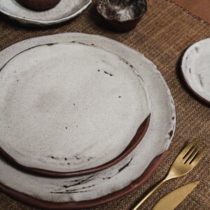 Pu&ograve; includere: Un set di piatti in ceramica bianca con bordi marroni, una piccola ciotola marrone e posate dorate su un sottopentola in tessuto marrone.