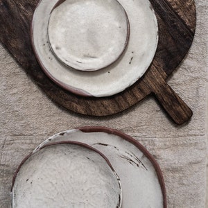 Pu&ograve; includere: Due set di piatti in ceramica bianca con bordi marroni. I piatti sono annidati e hanno un aspetto rustico e fatto a mano.