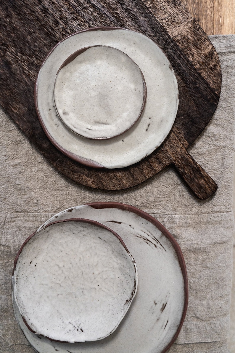 Pu&ograve; includere: Due set di piatti in ceramica bianca con bordi marroni. I piatti sono annidati e hanno un aspetto rustico e fatto a mano.