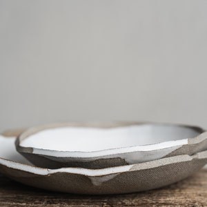 Pu&ograve; includere: Due ciotole in ceramica bianca con un esterno ruvido e testurizzato. Le ciotole sono impilate l'una sull'altra e sono appoggiate su un pezzo di legno invecchiato.