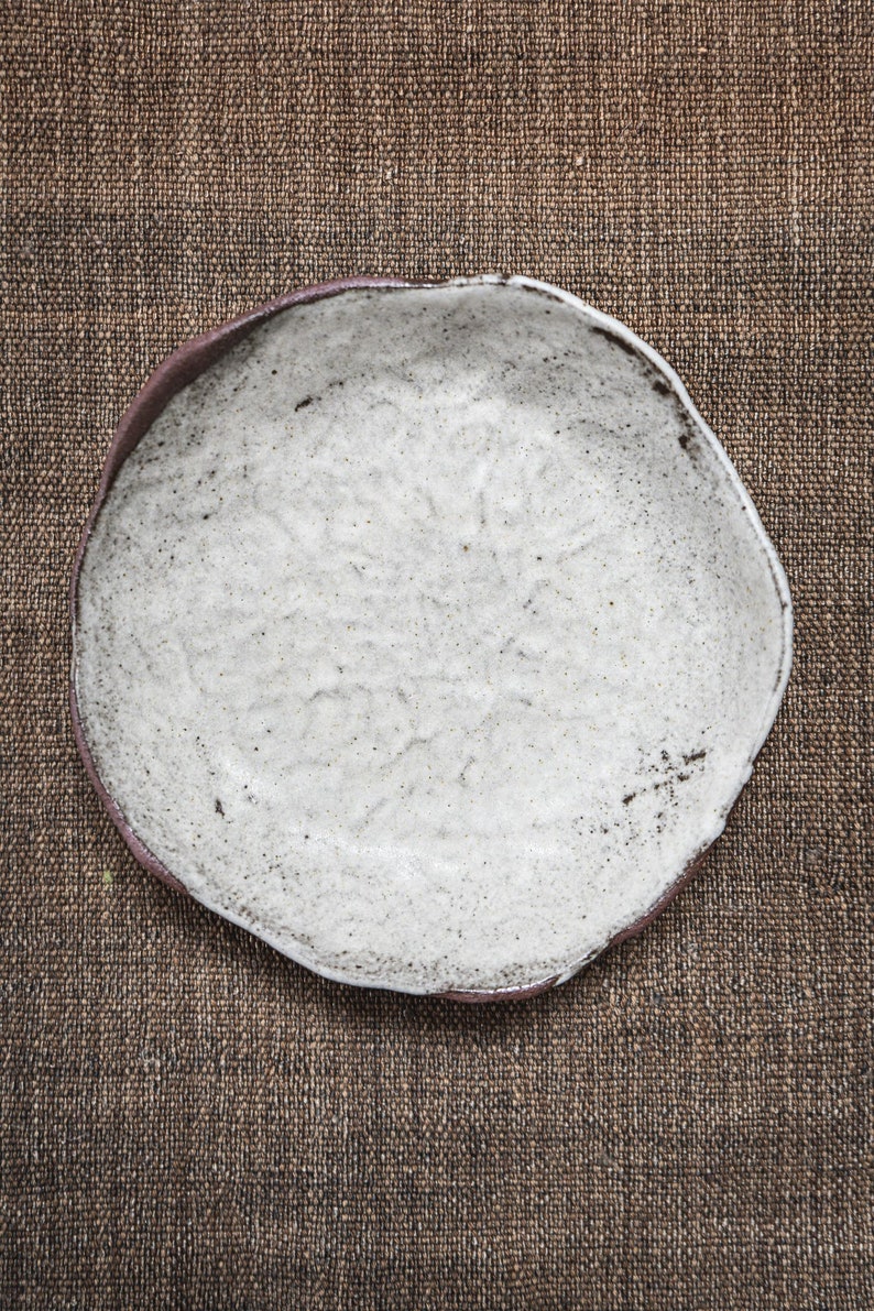 Pu&ograve; includere: Un piatto in ceramica bianca con una superficie testurizzata e una forma leggermente irregolare. Il piatto &egrave; appoggiato su uno sfondo di juta marrone.