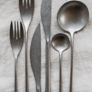 Op de afbeelding: Een set rustiek bestek, bestaande uit twee vorken, twee messen en twee lepels, gerangschikt op een wit linnen oppervlak. Het bestek heeft een matte, metallic afwerking en de gebruiksvoorwerpen vari&euml;ren in grootte en vorm.