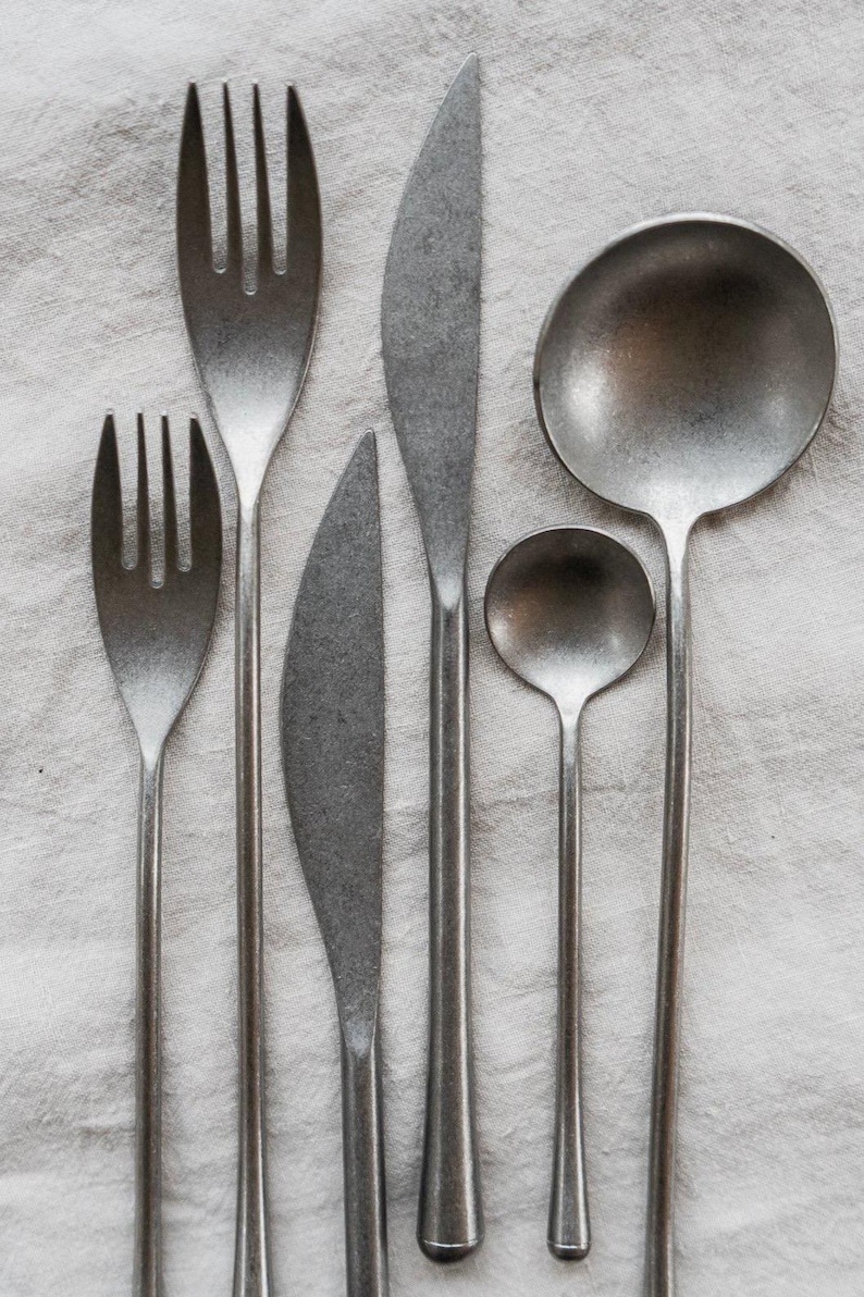 Op de afbeelding: Een set rustiek bestek, bestaande uit twee vorken, twee messen en twee lepels, gerangschikt op een wit linnen oppervlak. Het bestek heeft een matte, metallic afwerking en de gebruiksvoorwerpen vari&euml;ren in grootte en vorm.