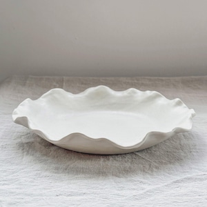 Può includere: Un piatto da portata in ceramica bianca con un bordo ondulato. Il piatto è appoggiato su una tovaglia di lino bianca.