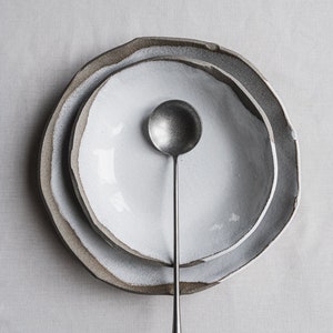 Pu&ograve; includere: Un set di due ciotole in ceramica bianca con un esterno ruvido e testurizzato. Le ciotole sono annidate l'una dentro l'altra e un cucchiaio d'argento &egrave; posto al centro delle ciotole.
