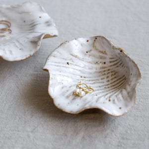 Puede incluir: Dos cuencos de cerámica blanca para joyas con diseño de hoja. Los cuencos tienen una textura moteada y un borde ligeramente ondulado. Uno contiene anillos de oro, el otro pendientes de oro con perlas.