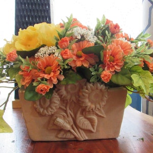 Op de afbeelding: Een bloemstuk in een terracotta pot met een zonnebloemdessin. Het arrangement bevat gele rozen, oranje en witte bloemen en groene klimop.