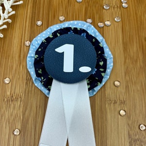 Könnte beinhalten: Eine blau-weiße Rosette mit der weißen Zahl "1.". Die Rosette hat ein Blumenmuster und ist an einem weißen Band befestigt. Das Wort "ander" ist auf einem kleinen weißen Etikett aufgedruckt.