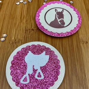 Könnte beinhalten: Zwei runde Untersetzer mit Pferdemotiv. Einer zeigt eine Pferdekopf-Silhouette in einem Kreis, der andere eine weiße Sattel-Silhouette. Beide haben einen pinkfarbenen Glitzerrahmen und einen cremefarbenen Hintergrund.