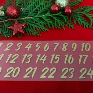 Könnte beinhalten: Eine transparente Plastikfolie mit goldenen Zahlen von 1 bis 24 darauf gedruckt. Die Zahlen sind in einer handschriftlichen Schriftart. Die Folie liegt auf einer roten Oberfläche mit einem grünen Tannenzweig und roten und goldenen Ornamenten.