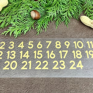 Könnte beinhalten: Eine transparente Plastikfolie mit goldenen Zahlen von 1 bis 24 darauf gedruckt. Die Zahlen sind in drei Reihen angeordnet. Die Folie liegt auf einer braunen Oberfläche.