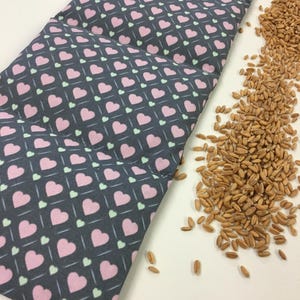 Könnte beinhalten: Ein graues Heizkissen aus Stoff mit einem Muster aus rosa und grünen Herzen. Das Kissen ist mit ungekochten Weizenkörnern gefüllt.