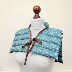 May include: A teal quilted wrap with a white star pattern, displayed on a mannequin. The wrap has a brown leather-like trim and ties in the front. The background is a plain off-white.