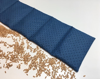 May include: A long, blue, polka-dotted fabric heating pad with four sections. The pad is filled with grains and is lying on a white surface with scattered grains.
