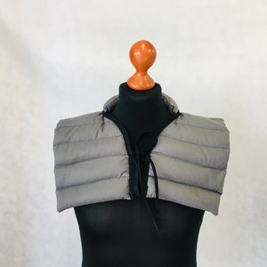 Könnte beinhalten: Ein grauer, gesteppter Nackenwärmer mit schwarzem Besatz und Kordelverschluss. Der Nackenwärmer wird auf einer schwarzen Schaufensterpuppe präsentiert, die sein Design und seine Form zeigt. Der Artikel soll Wärme und Stil bieten.