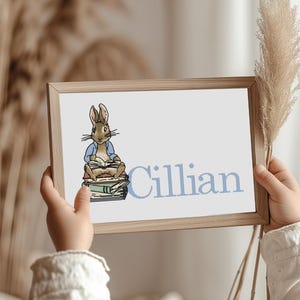 Op de afbeelding: Een ingelijste print met een aquarel illustratie van een konijn dat leest op een stapel boeken. De naam "Cillian" is in blauw gedrukt. De lijst is lichtbruin.