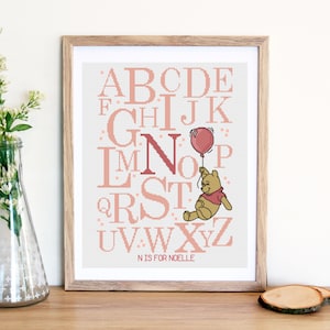 May include: A framed cross-stitch alphabet chart with a pink and white color scheme. The letter N is highlighted with a pink Winnie the Pooh character holding a pink balloon. The text below the alphabet reads "N is for Noelle."