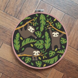 以下が含まれることがあります： ジャングルを背景に、枝にぶら下がる3匹のナマケモノを描いた刺繍のフープアート。ナマケモノは茶色で黒い目をしていて、緑の葉とツルに囲まれています。