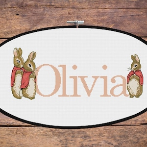 May include: A white oval-shaped canvas with a black frame, featuring two brown rabbits wearing red jackets and the name "Olivia" in pink lettering.