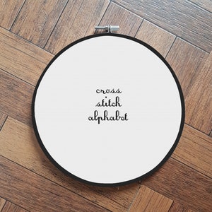 May include: White embroidery hoop with black text that reads "cross stitch alphabet".