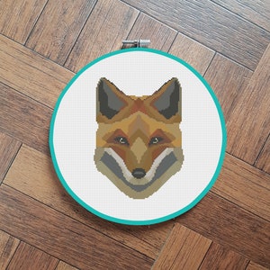 Könnte beinhalten: Ein Kreuzstich-Stickrahmen mit einem geometrischen Fuchs-Design. Der Fuchs ist braun, orange und grau mit einer weißen Schnauze.