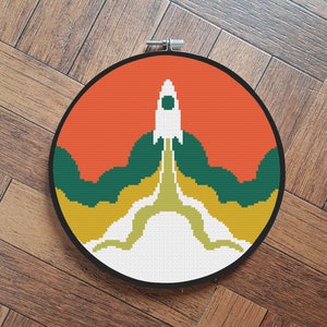 May include: A round embroidery hoop with a colorful design of a rocket ship launching into a red, yellow, and green sky.
