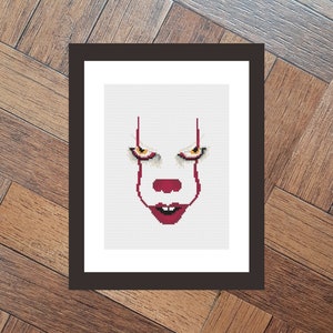 May include: Framed cross-stitch artwork depicting a clown's face with red details and yellow eyes. The design is set against a white backdrop, enclosed within a dark brown frame. The clown's face is a minimalist design.
