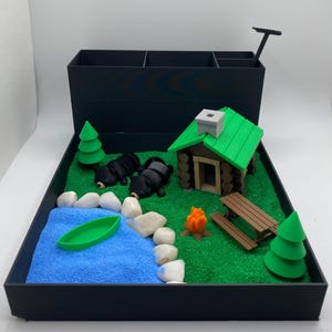 May include: A black desktop organizer with a miniature forest scene. The scene includes a small cabin, two black bears, a campfire, a picnic table, a lake, and two green pine trees. The organizer has a black base and a removable tray with a green grass-like surface.